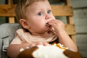 A small child in a highchair snacks on food with messy fingers and an expressive face.