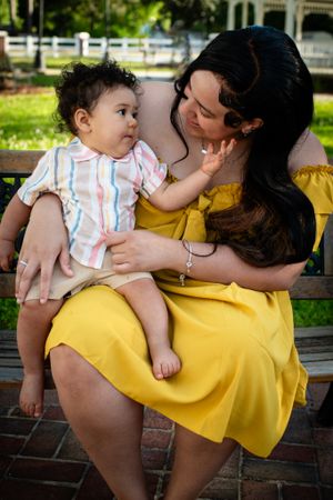 Mother in yellow dress sitting outdoors with baby on park bench.
