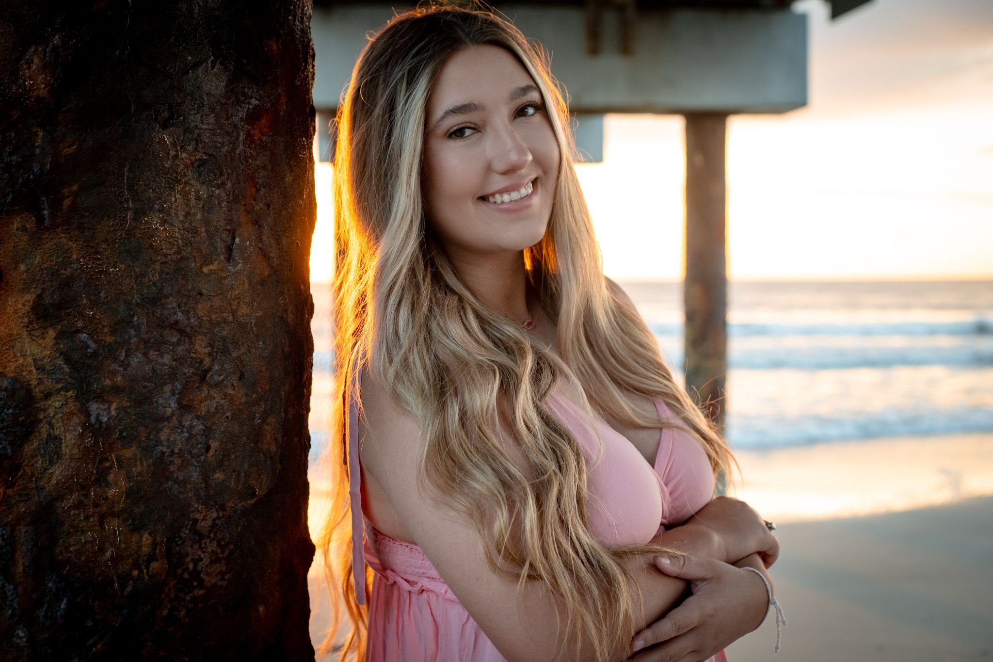 Portrait of someone in a pink top smiling at sunset by a beach pier.