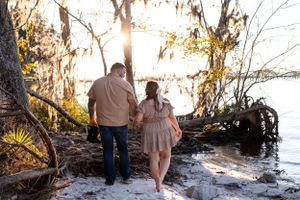 A couple walks along a beach shoreline at sunset among mossy trees.