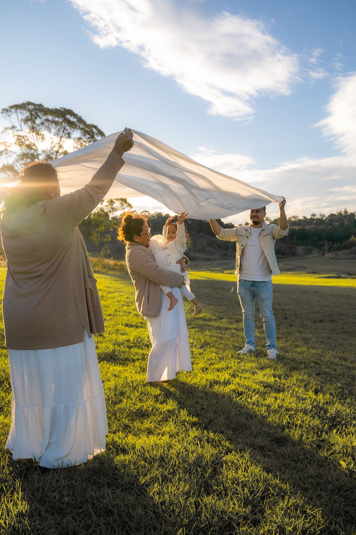 Mother and father hold a white sheet while the grandma and toddler dance under it at a photo shoot in The Dairy, Western Sydney Parklands.