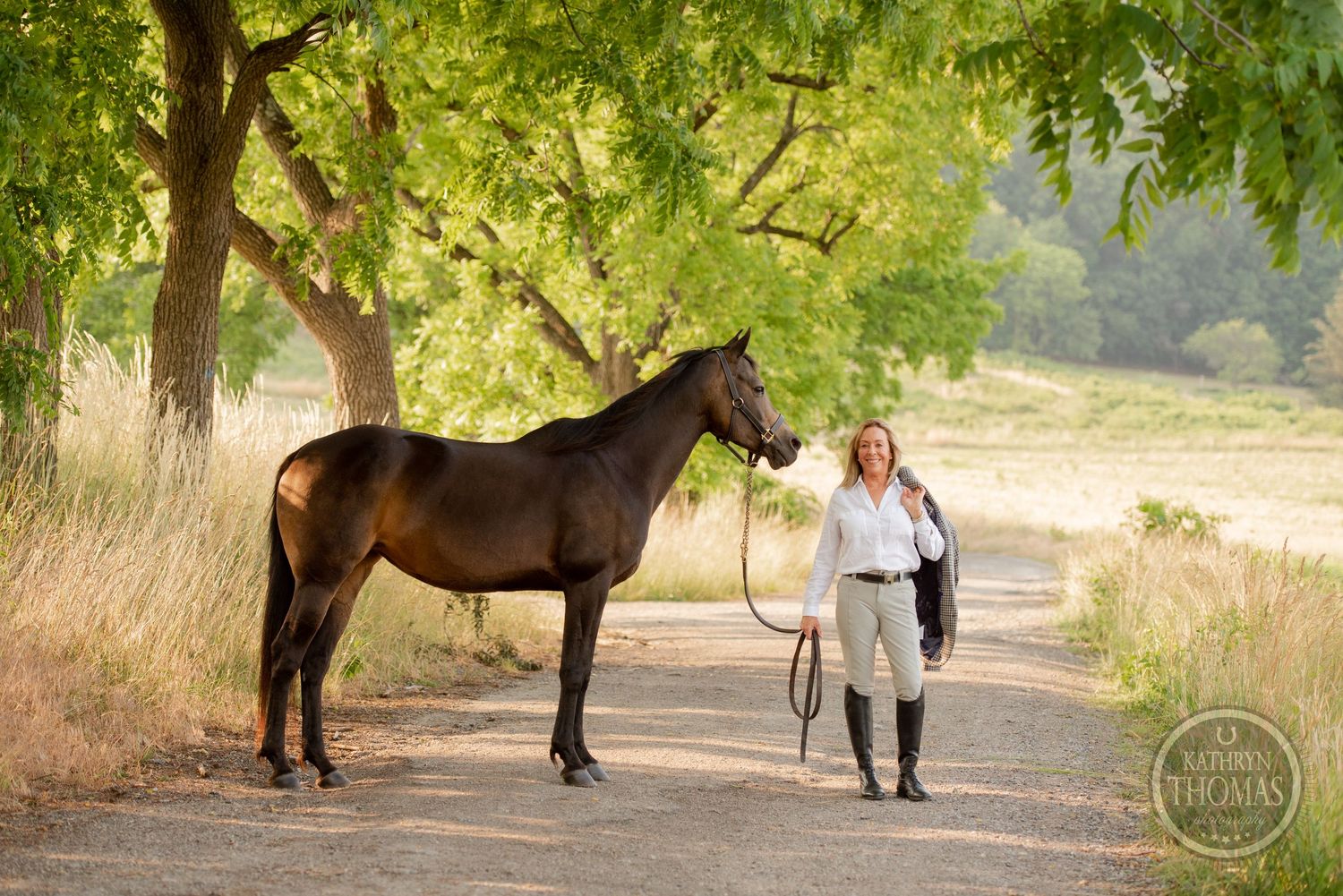 A rider poses with their majestic dark horse on a sunlit country path under green trees.
