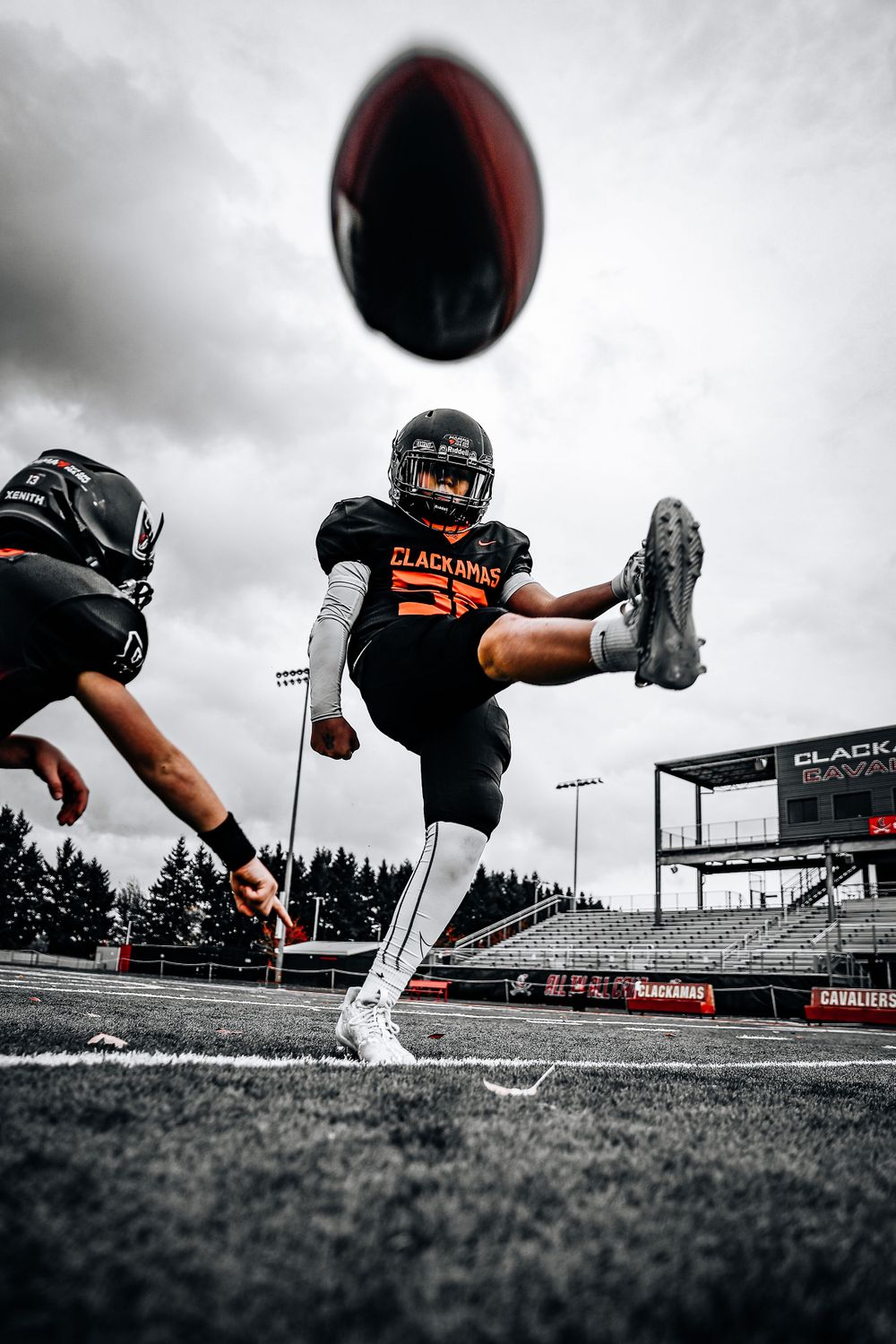 Football player in black uniform kicking ball against cloudy sky on stadium field.