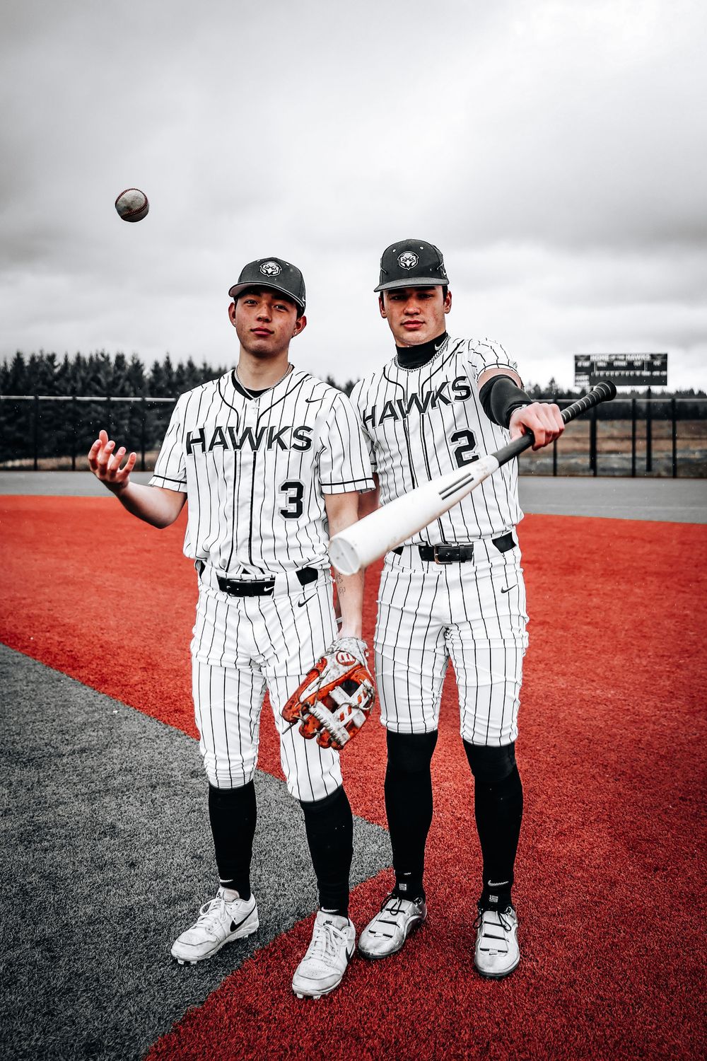 Two baseball players in pinstriped Hawks uniforms pose together on a red turf field with a baseball in the air.