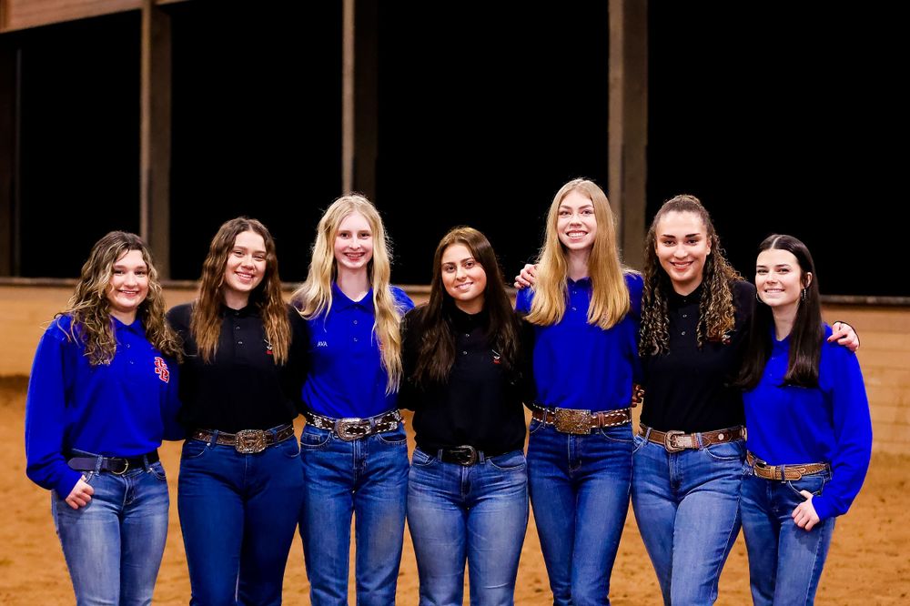 Team photo of equestrians wearing matching blue tops and jeans in indoor arena.