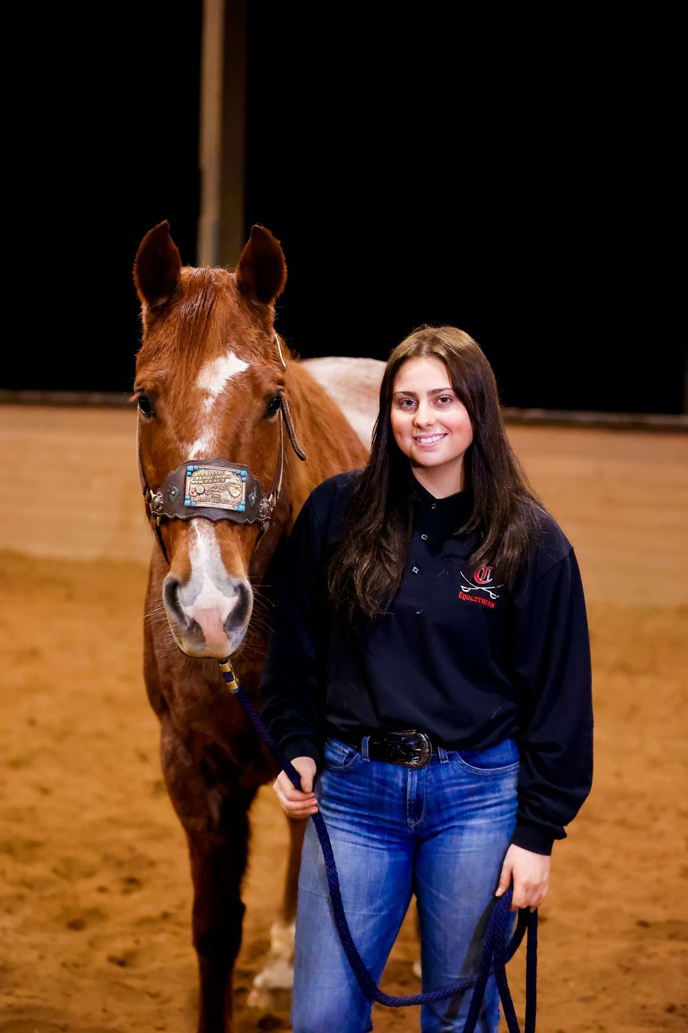 Student equestrian standing with brown horse in indoor arena wearing navy sweatshirt and jeans.