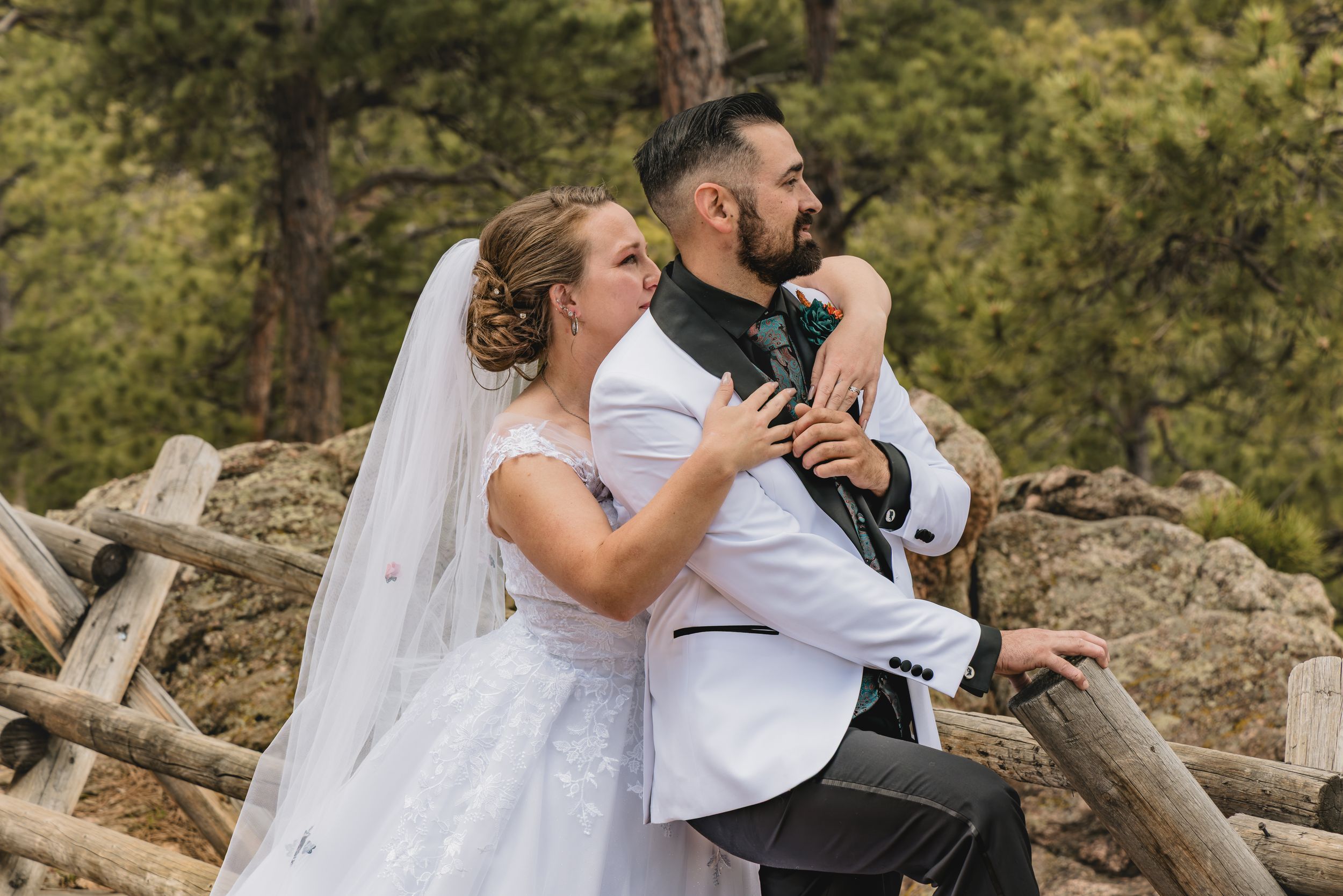 A couple in wedding attire share a tender moment on a rocky mountain overlook surrounded by pine trees.