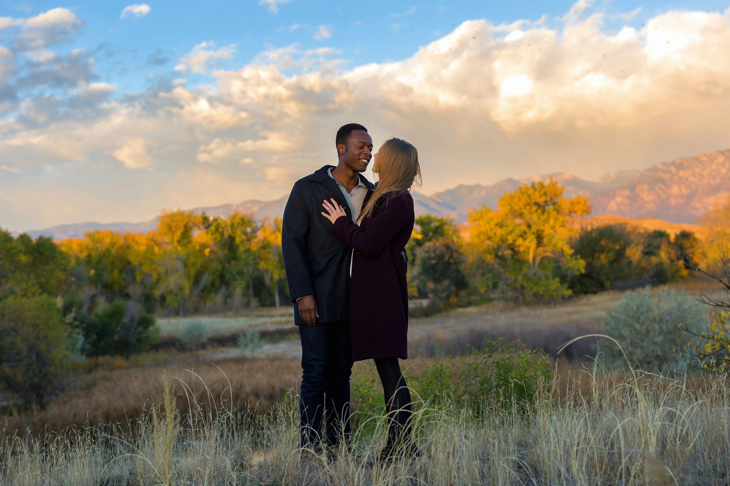 Couple sharing an intimate moment at sunset with mountains and golden trees in background.