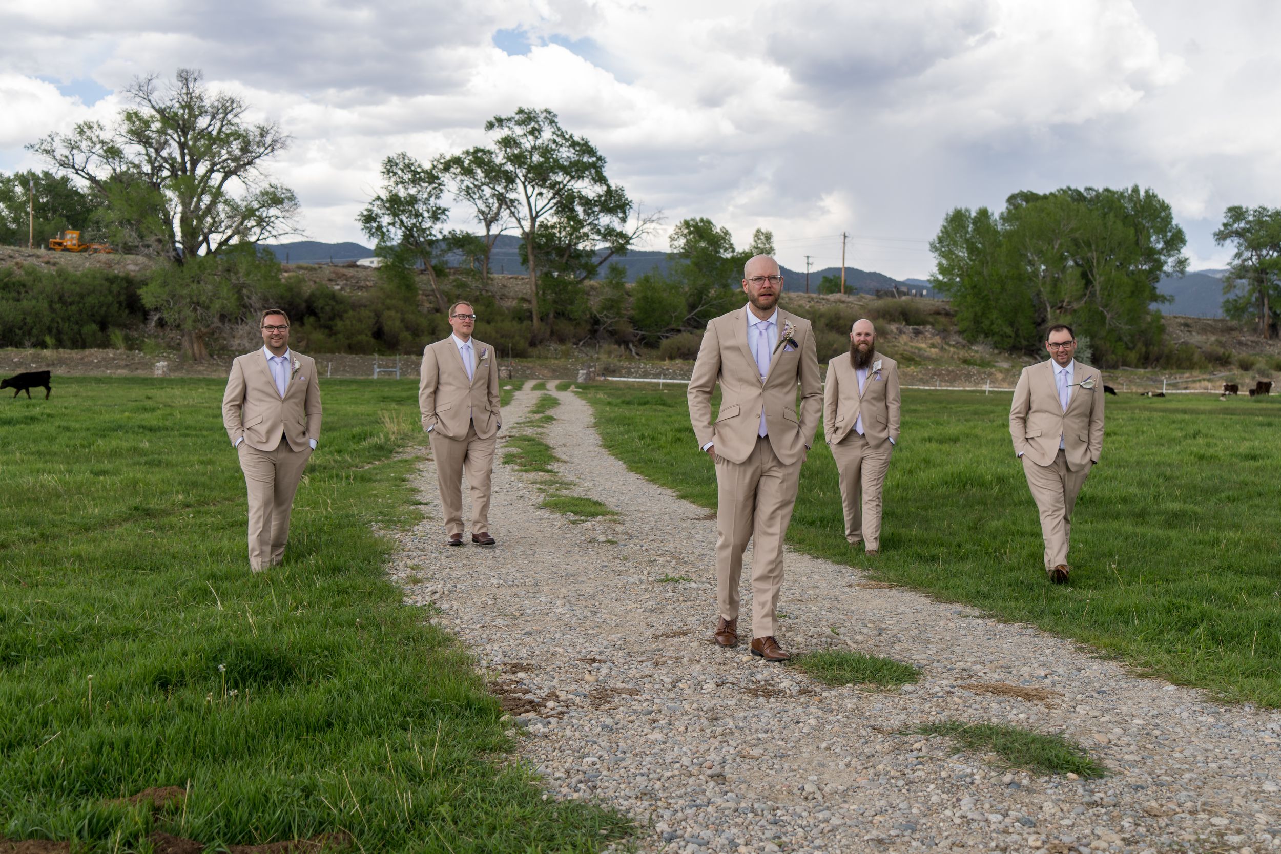 Five figures in beige suits walk along a gravel path through a green countryside landscape under cloudy skies.