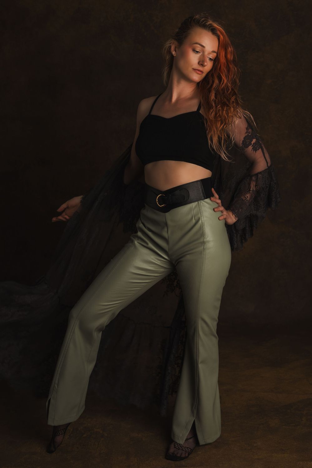 Artistic fashion portrait with black lace top, olive pants and moody lighting against dark brown background.