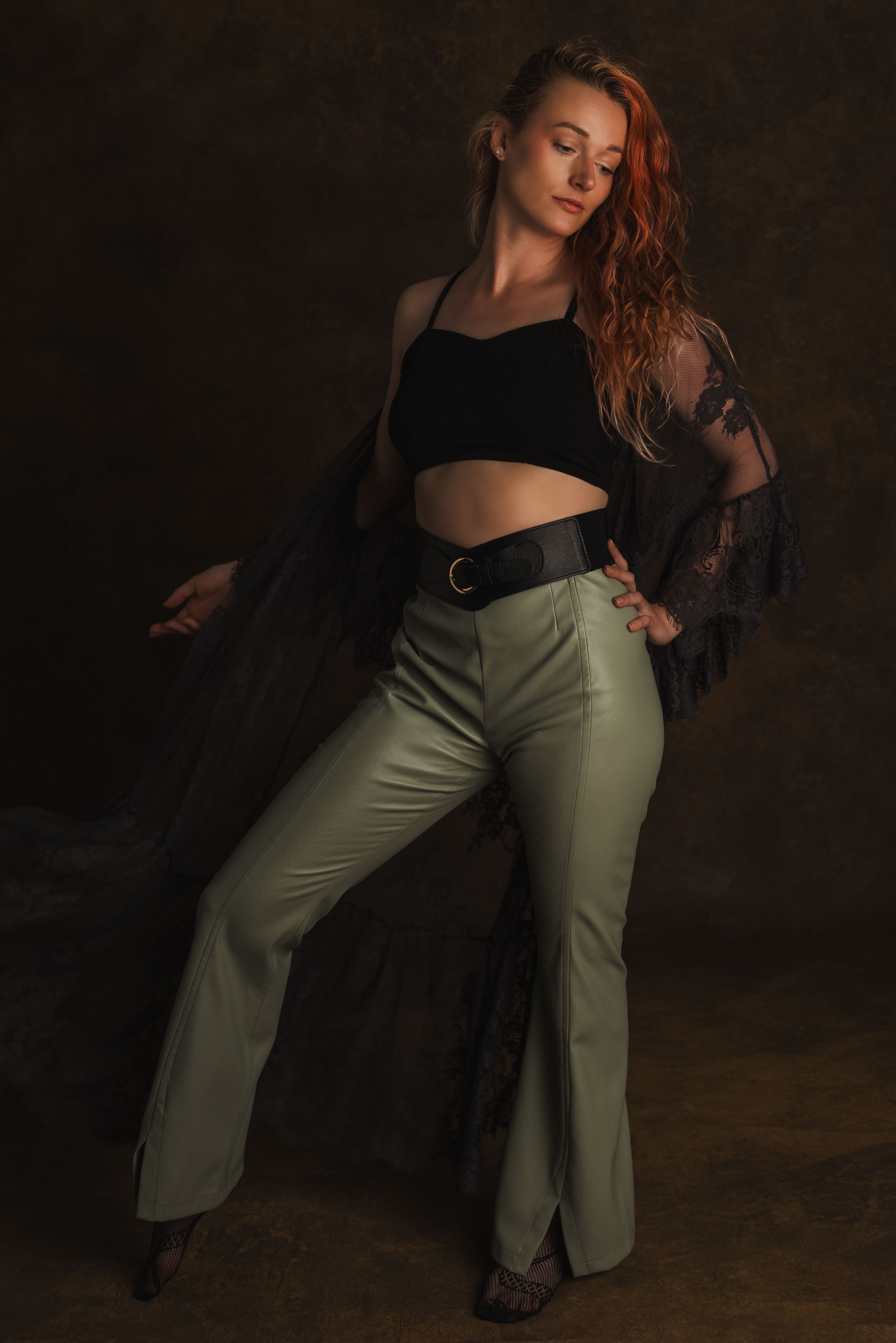 Artistic fashion portrait with black lace top, olive pants and moody lighting against dark brown background.