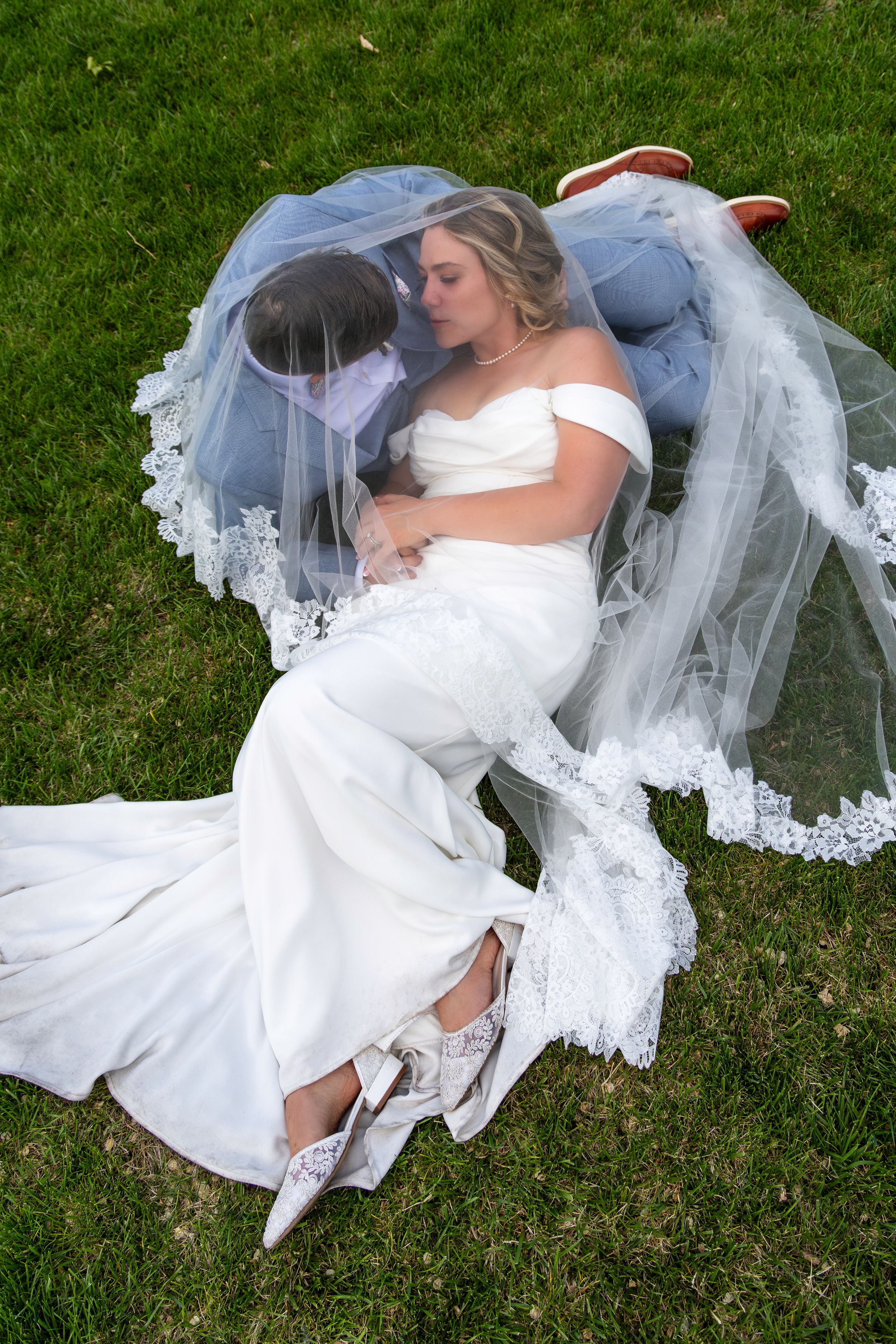 A person in a white wedding dress and veil lays on green grass during an outdoor wedding photo session.
