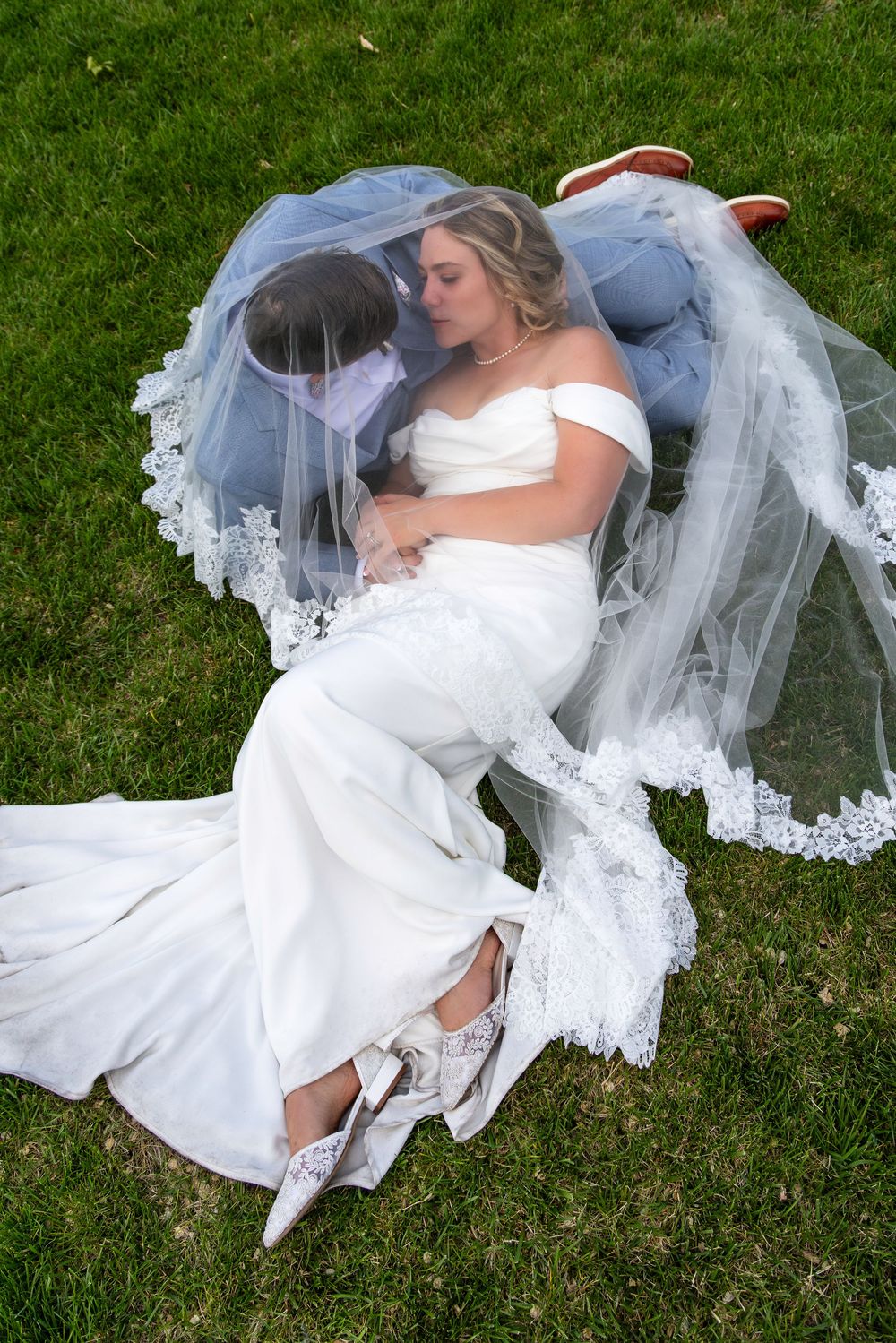 A person in a white wedding dress and veil lays on green grass during an outdoor wedding photo session.