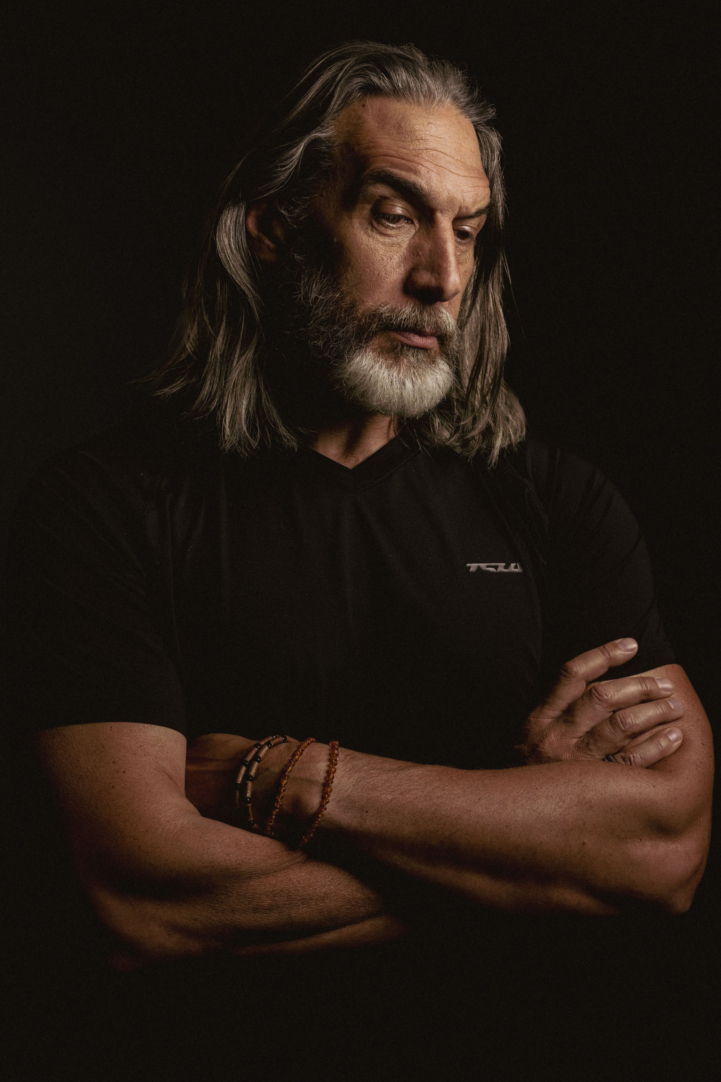Dramatic portrait of person with long gray hair and beard wearing black shirt against dark background.