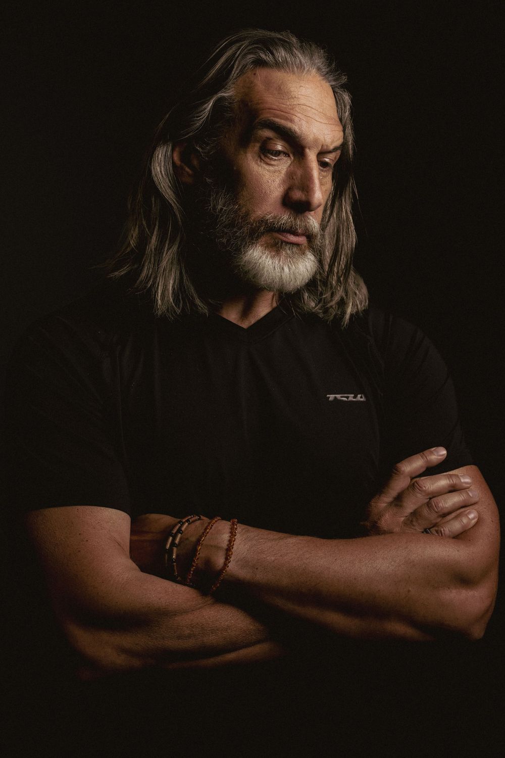 Dramatic portrait of person with long gray hair and beard wearing black shirt against dark background.