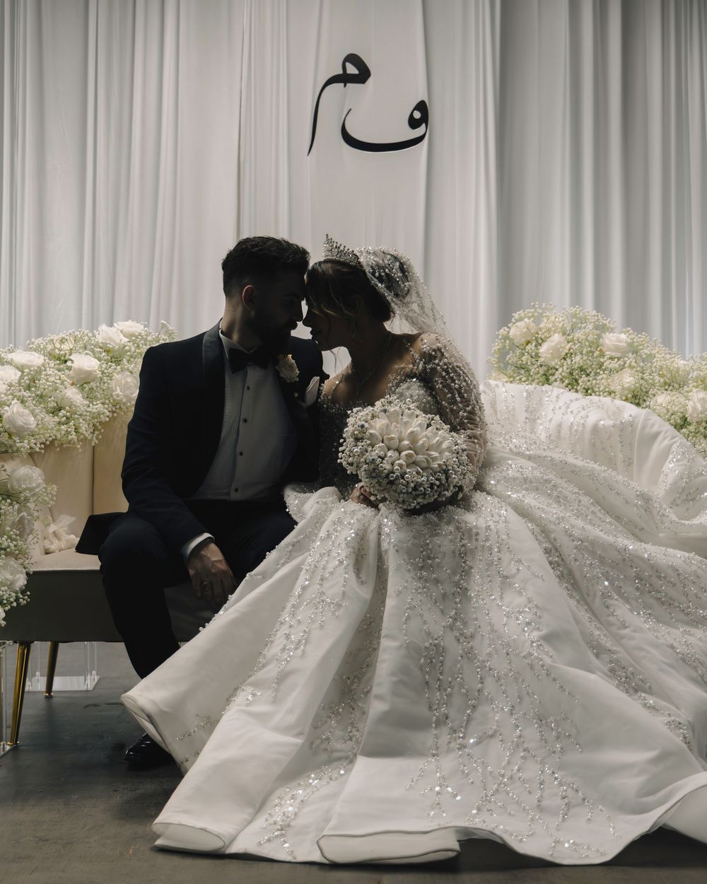 A romantic wedding moment with couple sharing an intimate pose against white floral decor and Arabic calligraphy on the wall.