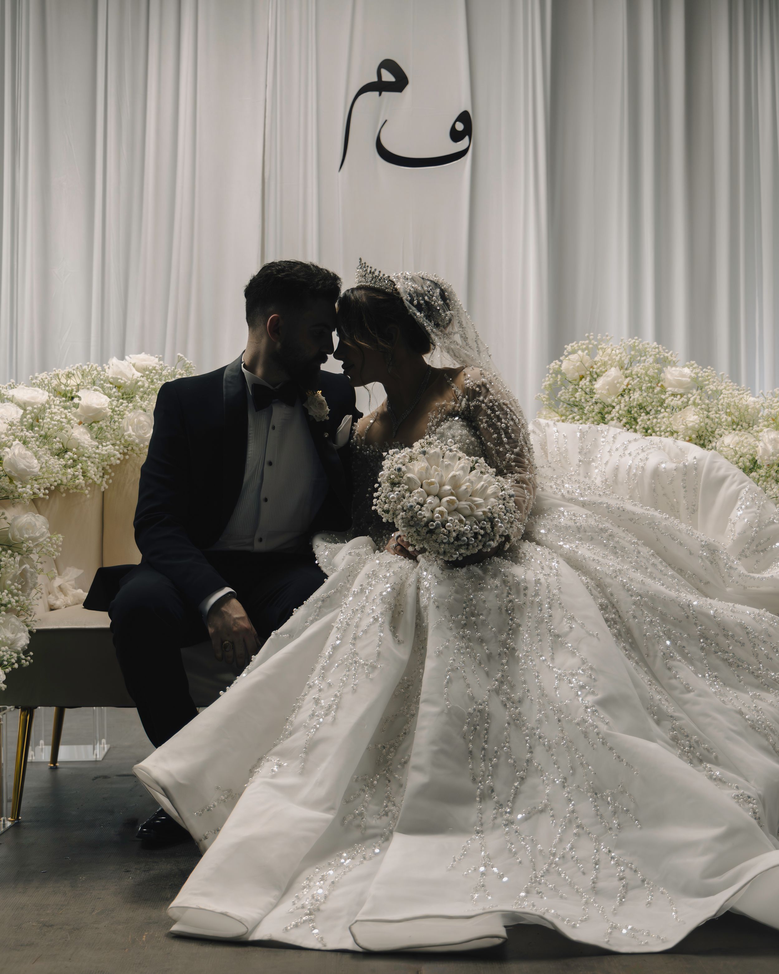 A romantic wedding moment with couple sharing an intimate pose against white floral decor and Arabic calligraphy on the wall.