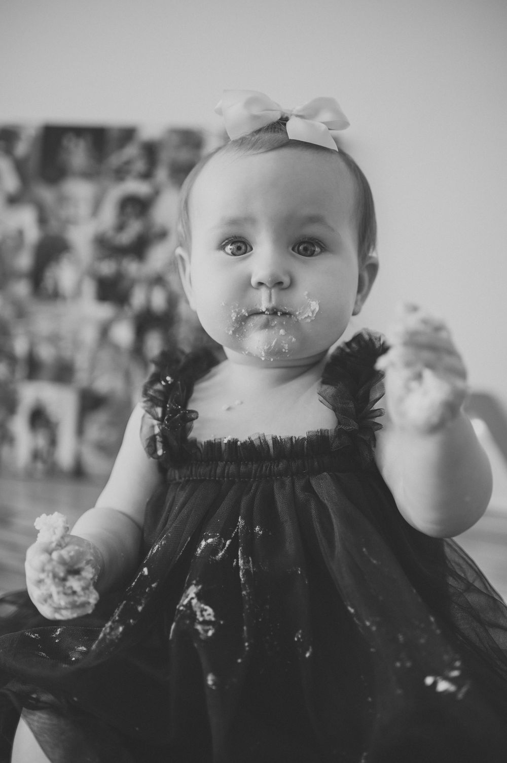 Black and white photo of baby girl with cake on face and hands wearing black dress with bow headband.