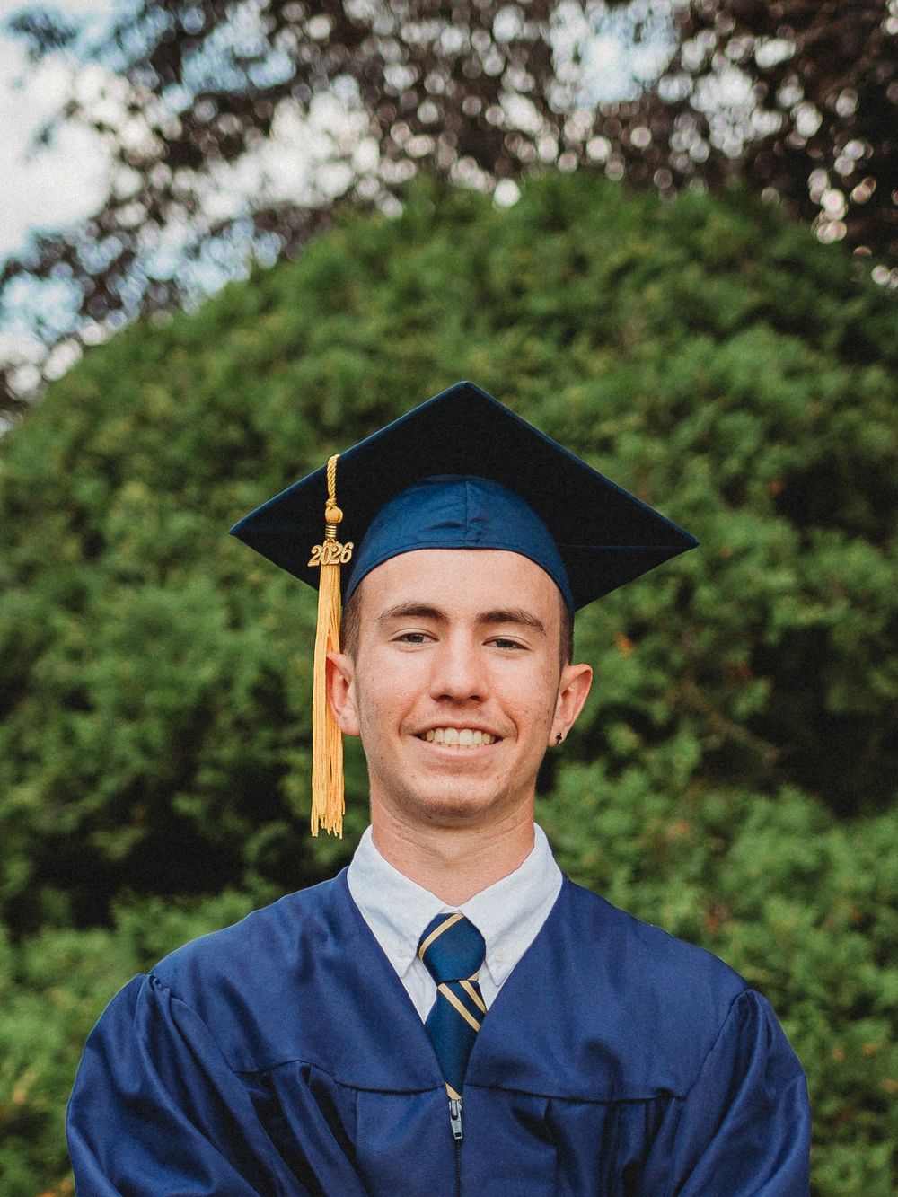 Happy young male graduate wearing navy blue cap and gown with gold tassel outdoors.