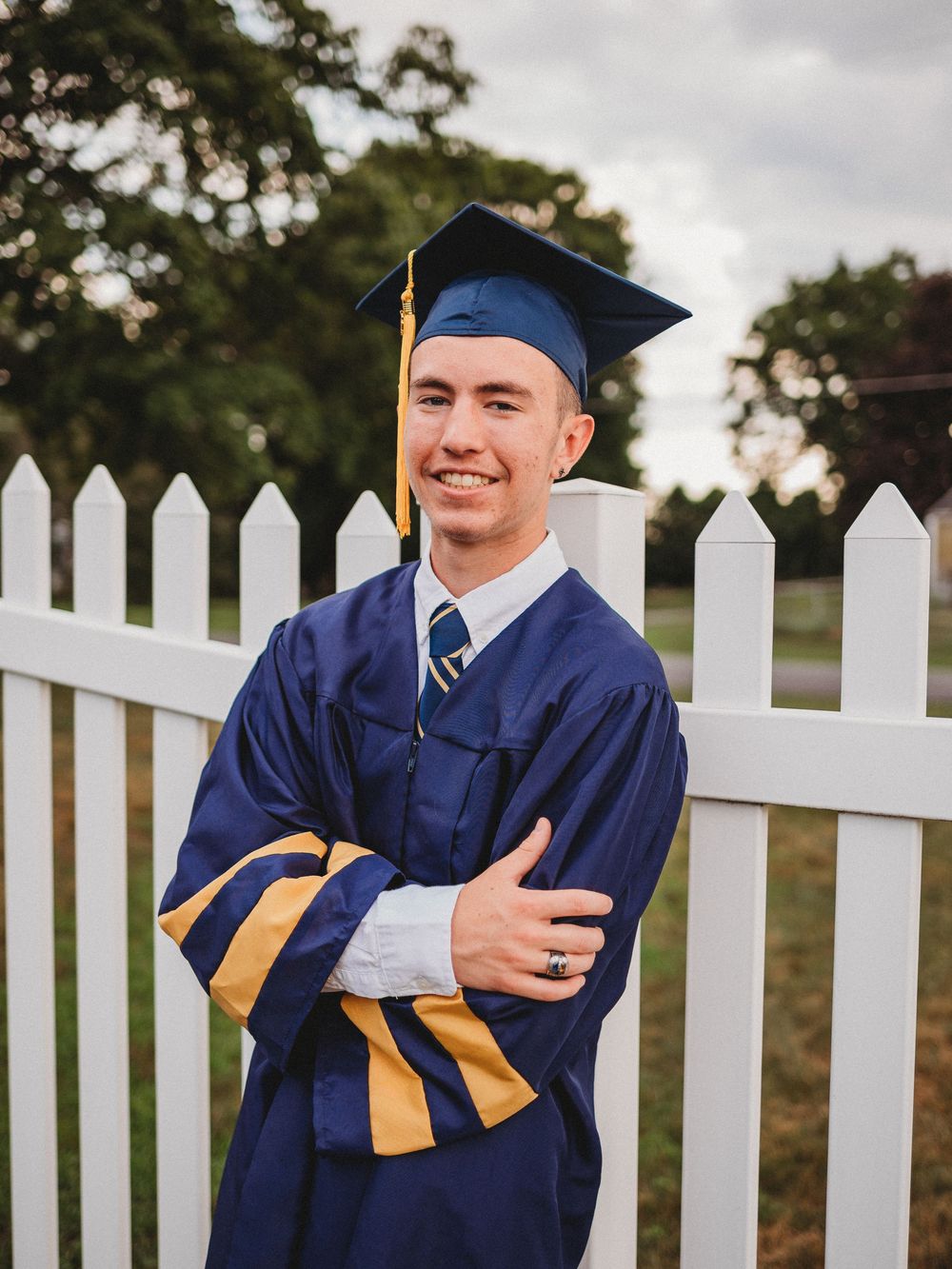 Smiling male graduate in navy blue and gold cap and gown posing by a white picket fence.
