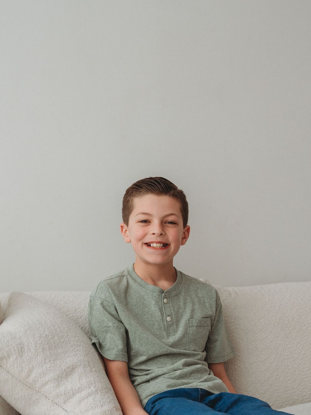 Smiling young boy in green henley shirt sitting on a white couch portrait photo.
