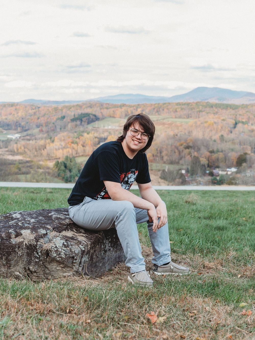 Teen in black tee sits on log with autumn valley landscape behind.