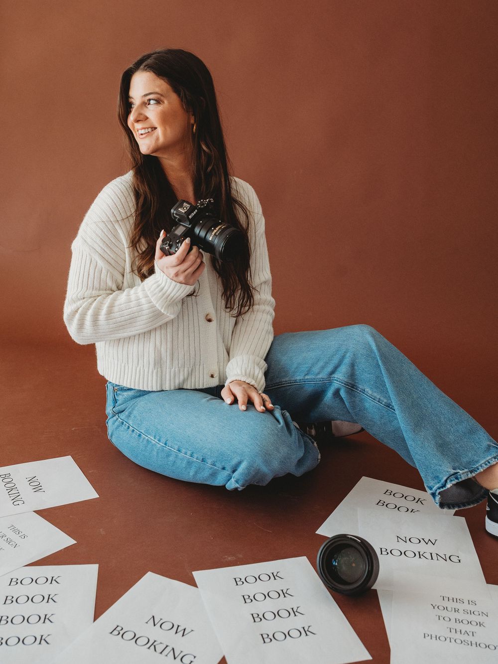 Female photographer sitting on floor with DSLR camera surrounded by booking promotional papers on brown background.