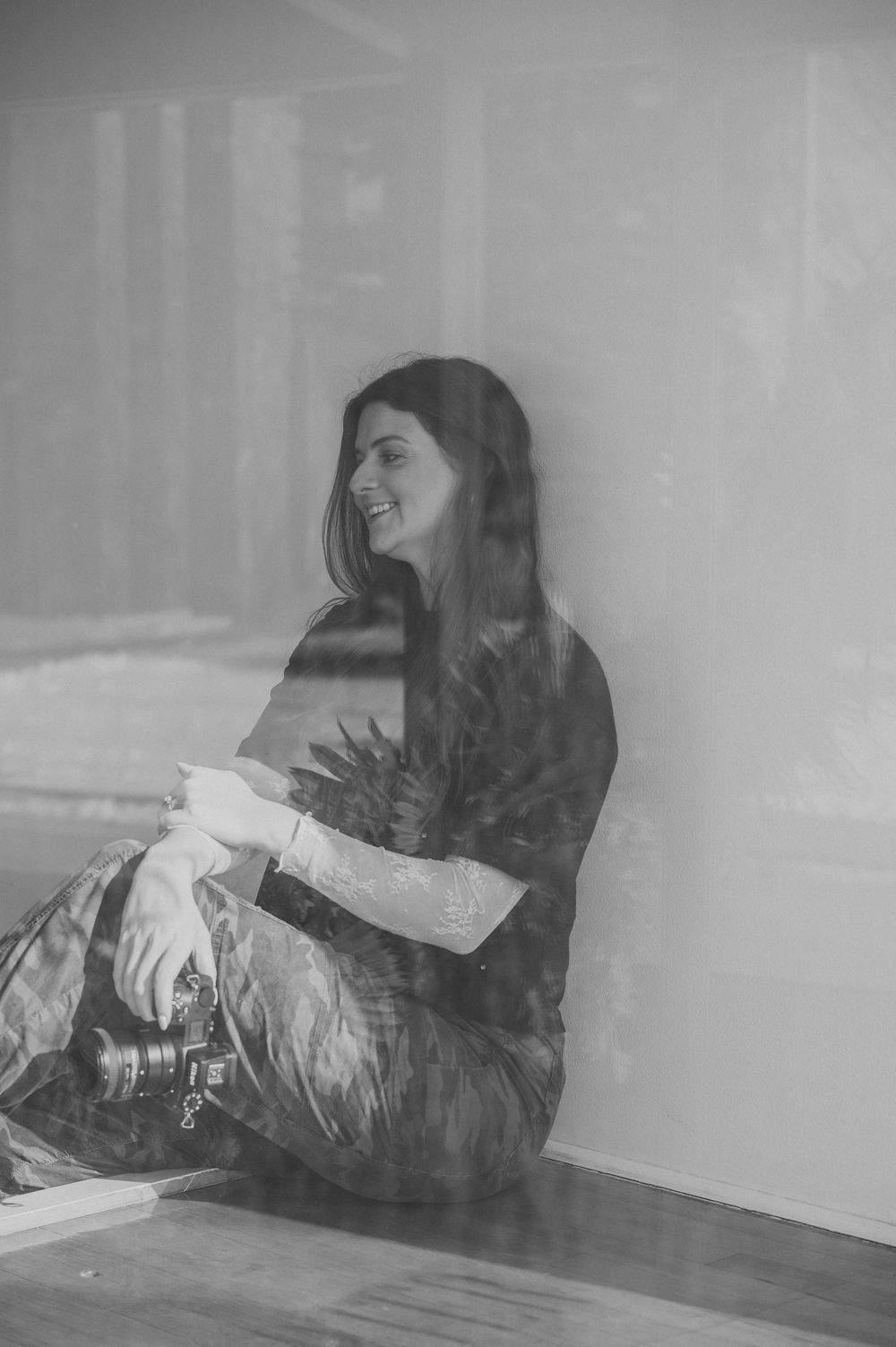 Black and white double exposure portrait of smiling woman with long dark hair holding camera indoors.