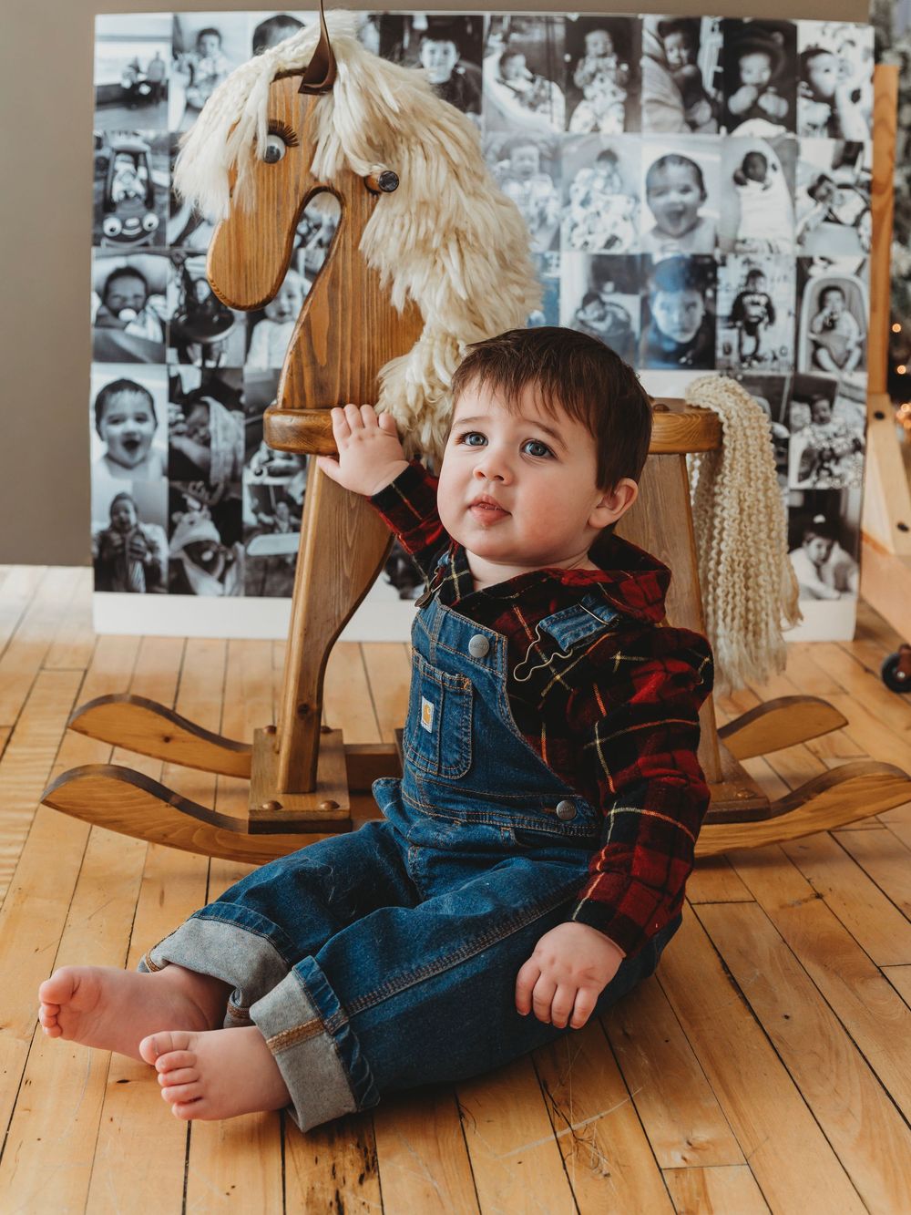 Cute toddler boy in denim overalls sitting beside a wooden rocking horse on hardwood floor.