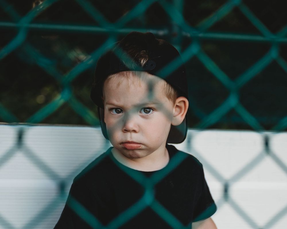 Young boy in black cap and shirt looking through chain-link fence with pouty expression.