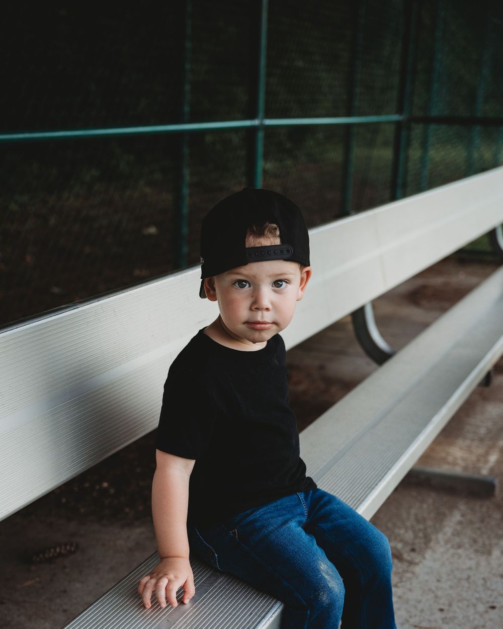 Toddler boy in black cap and shirt sitting on metal bleachers at baseball field.
