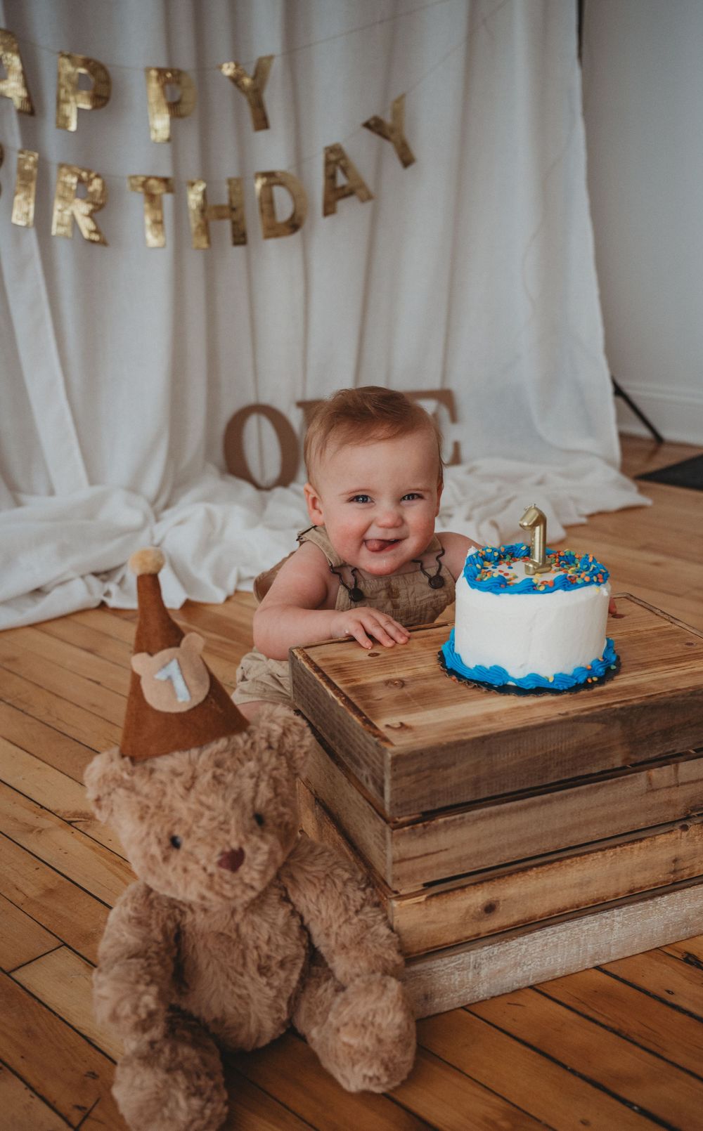 Smiling baby celebrates first birthday with blue and white smash cake beside teddy bear.