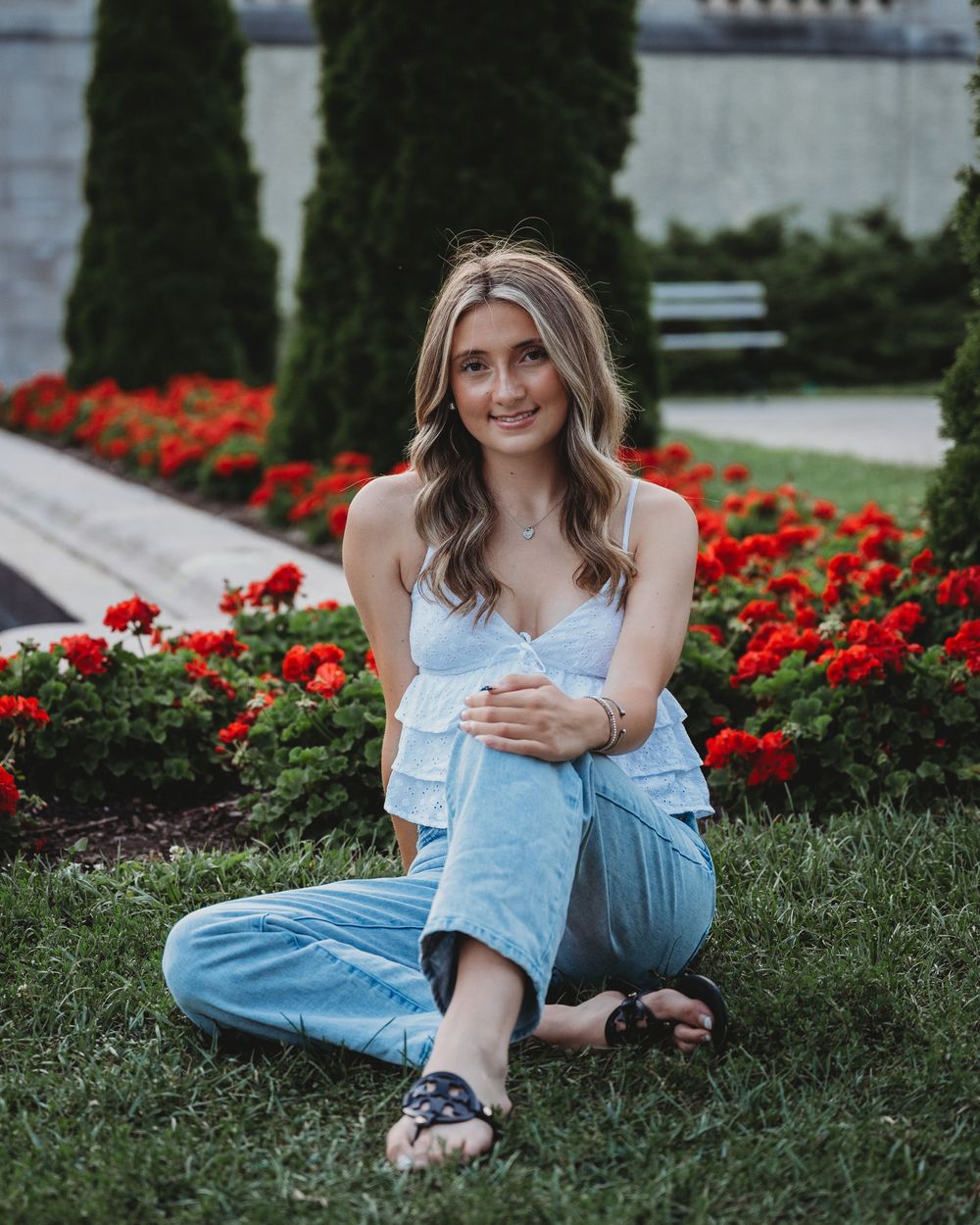 Smiling young woman in a white ruffled top and jeans sitting on grass near a red flower garden.