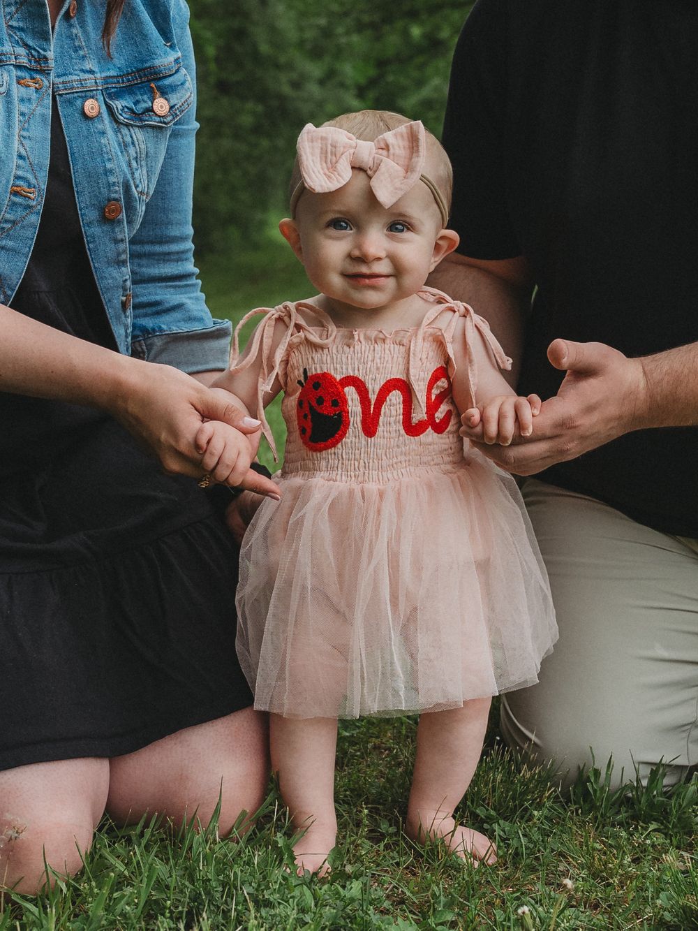Baby girl in pink tutu dress with 'one' embroidery stands with parents' support during first birthday photos.