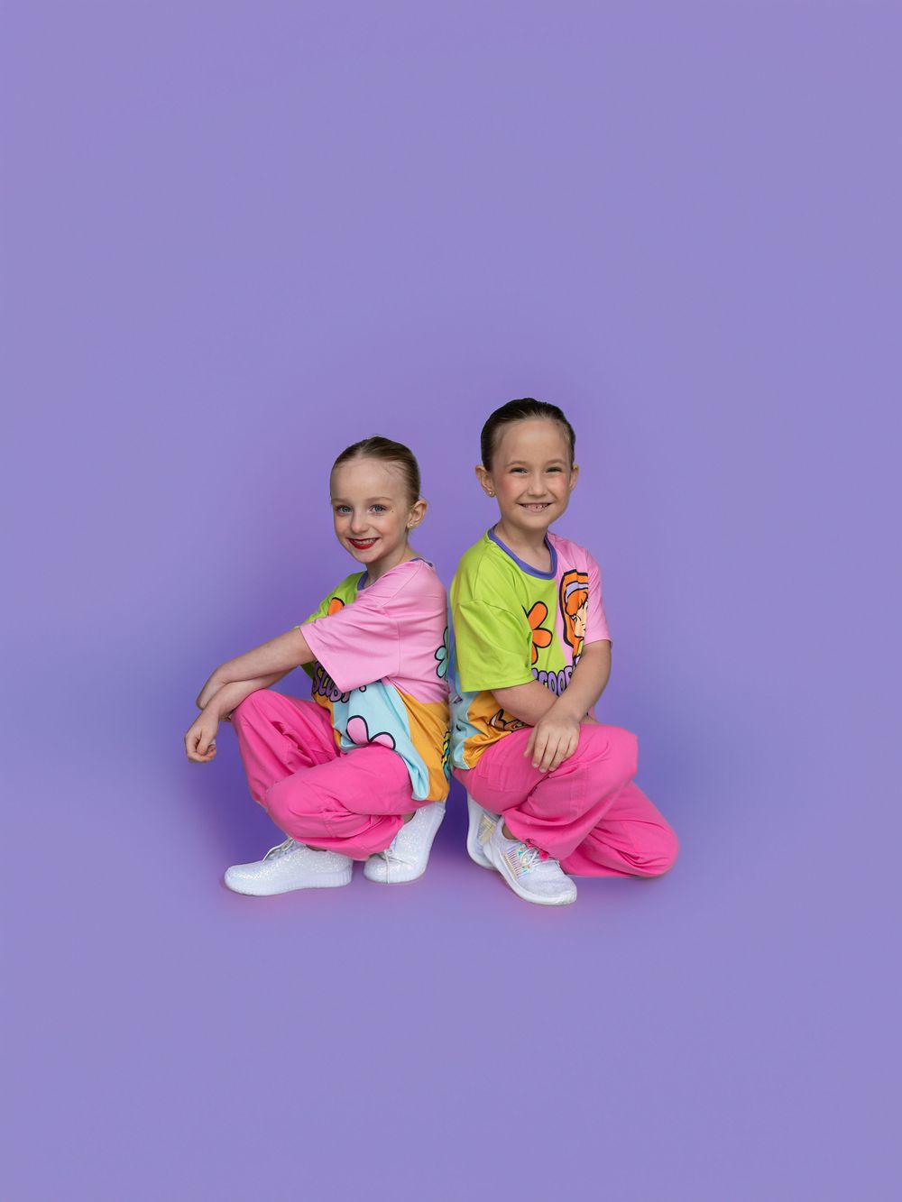 Two smiling girls in matching pink pants and colorful tops pose back-to-back on purple.