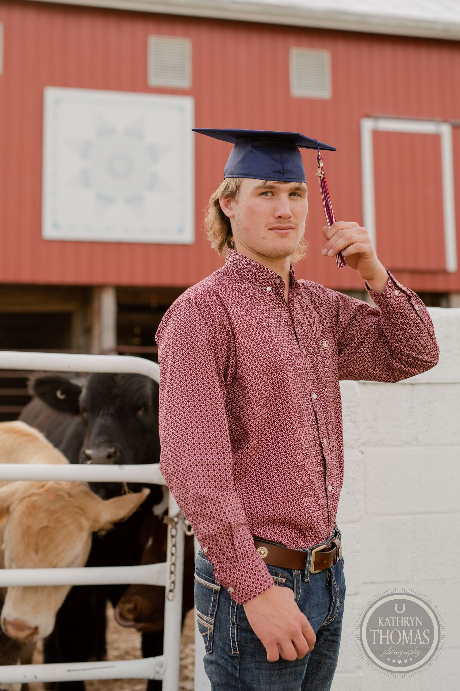 Person in red patterned shirt wearing graduation cap poses near barn with cattle visible.