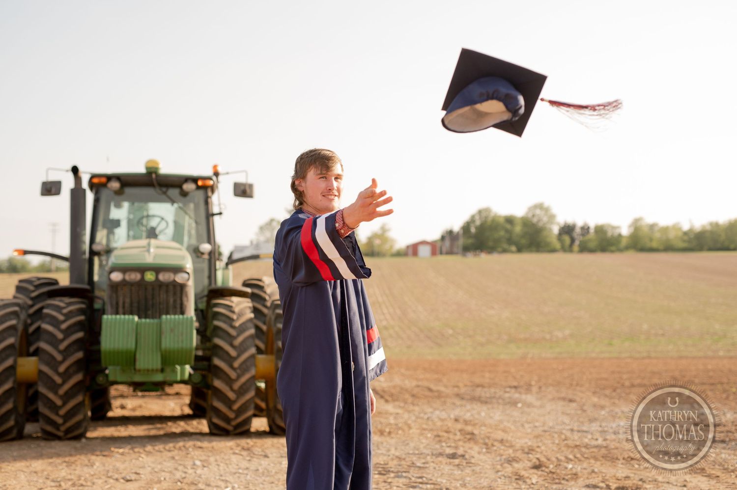 Graduate in cap and gown tosses mortarboard in air by tractor.