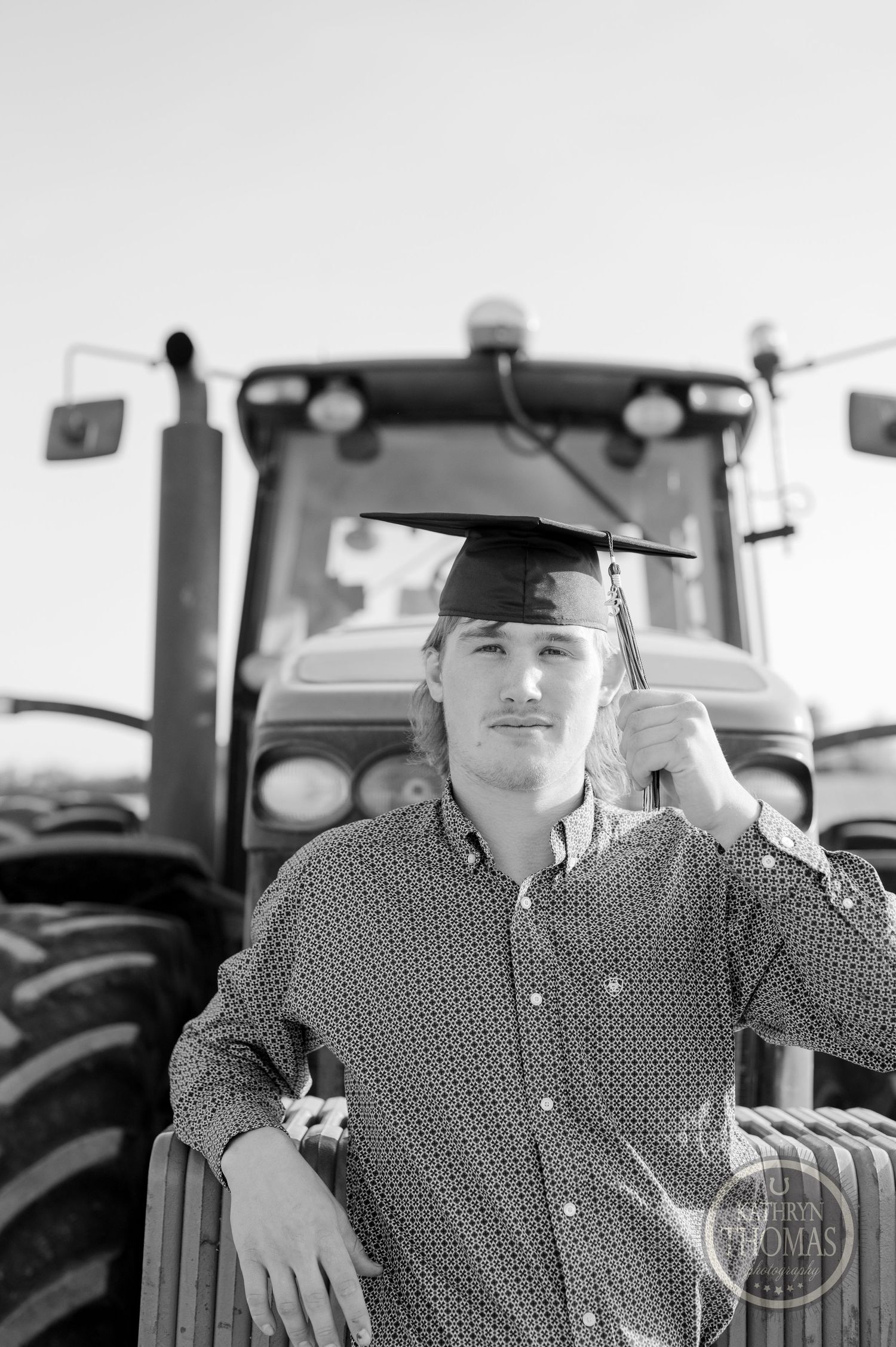 Black and white portrait of graduate in plaid shirt standing by farm tractor.