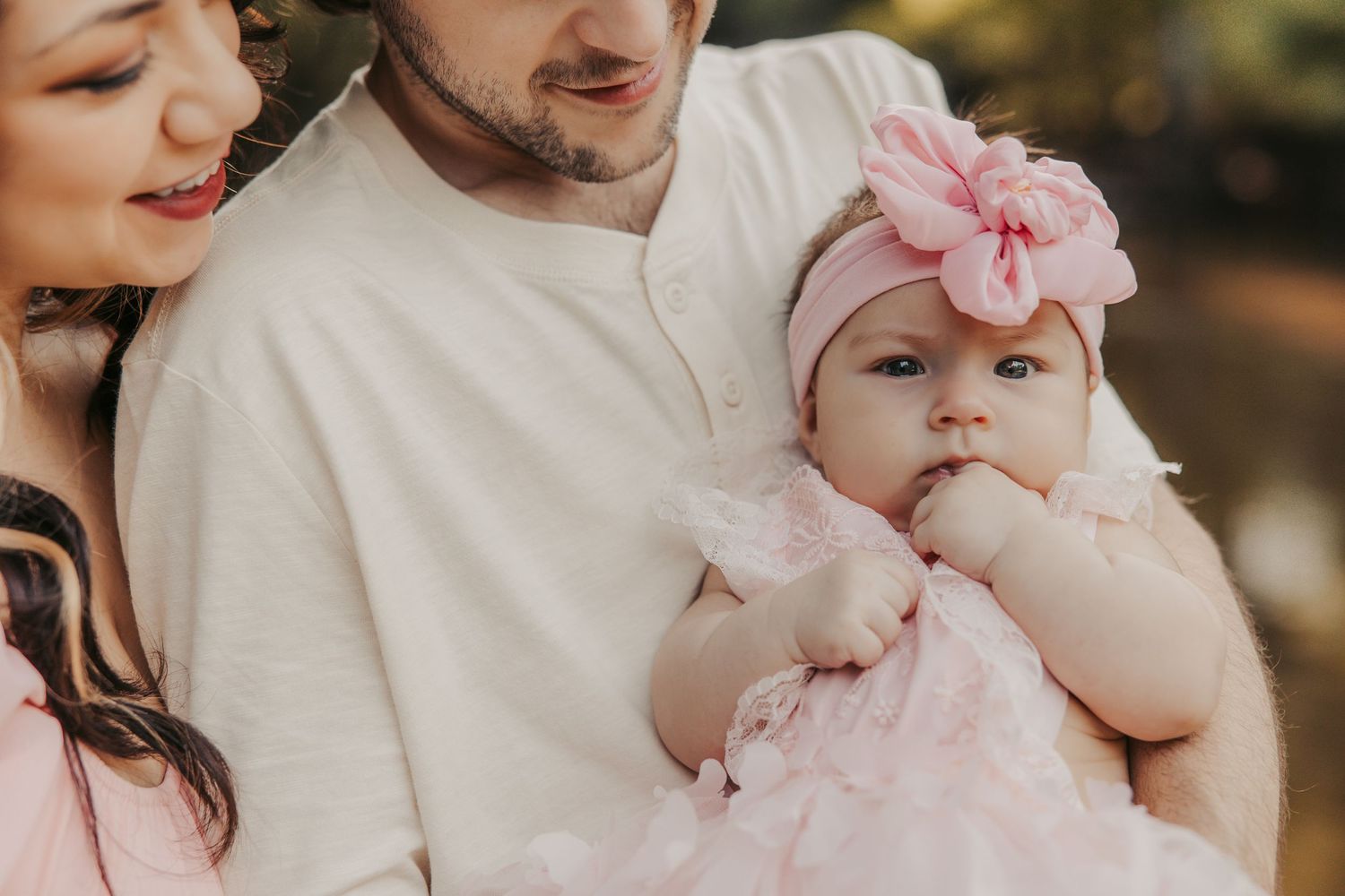 Baby in pink dress and floral headband being held by parents outdoors.