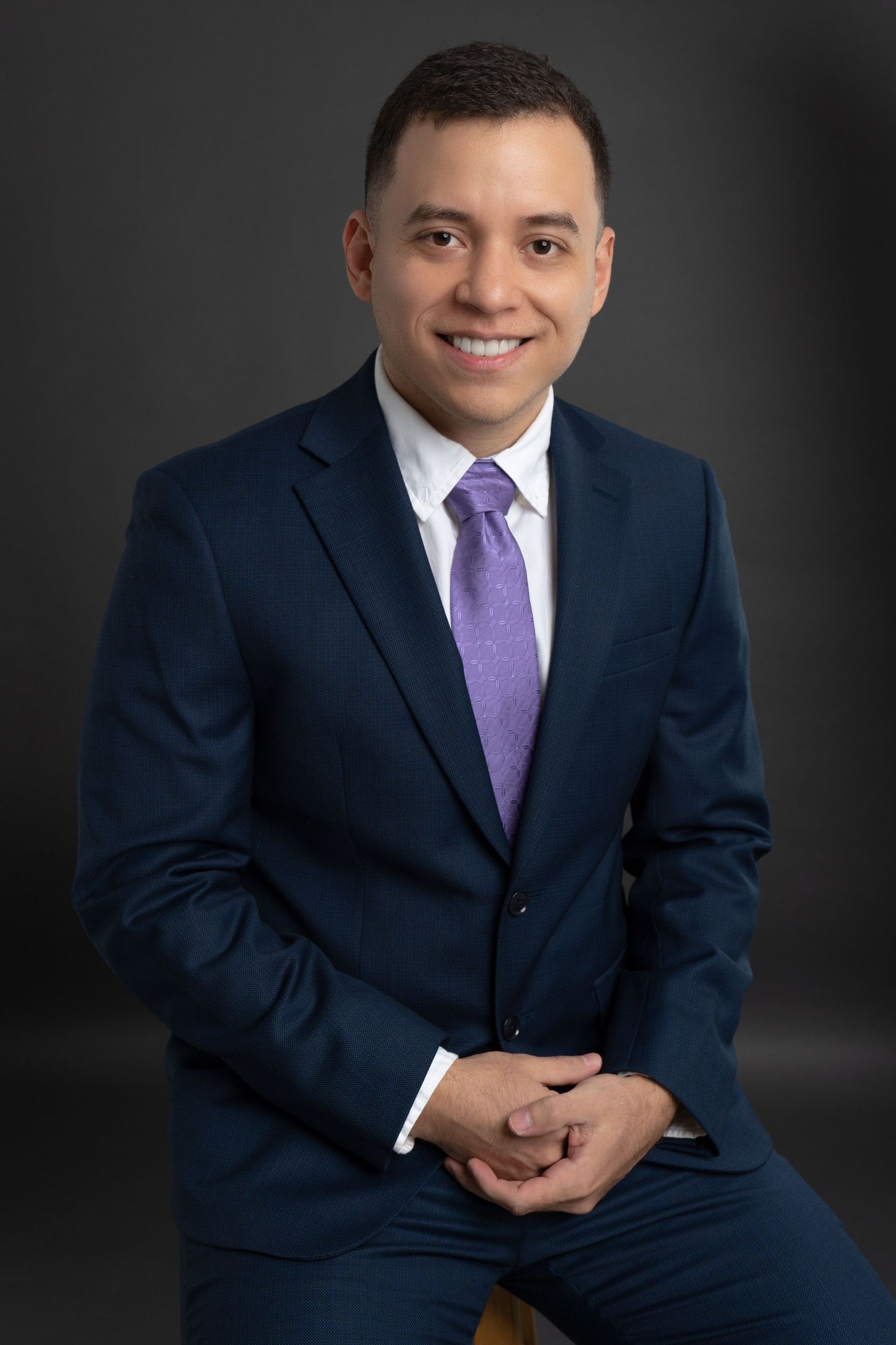 Professional headshot of person in navy suit and purple tie against dark background.