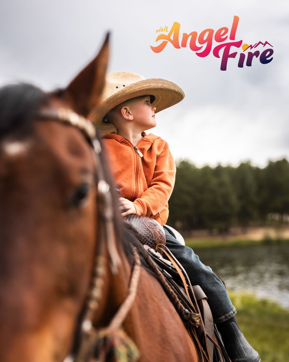 Young child in cowboy hat riding a brown horse at Angel Fire resort with trees and pond in background.