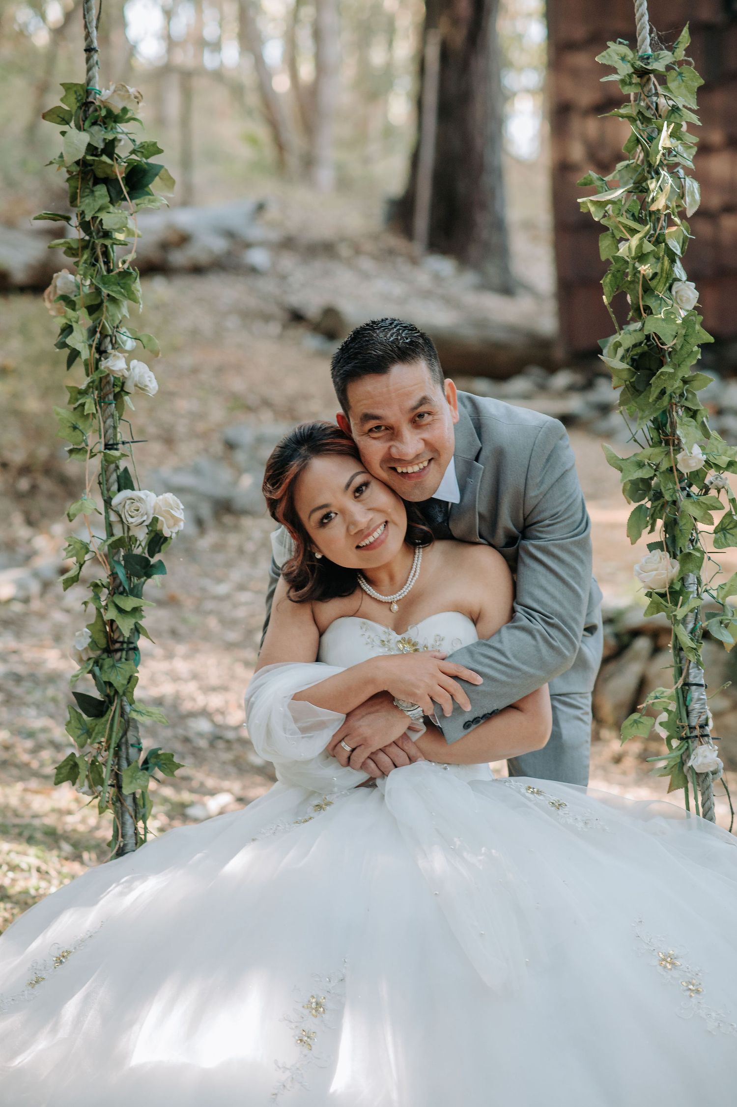 Newlyweds pose between greenery-draped swings in an enchanting forest setting during their wedding photoshoot.