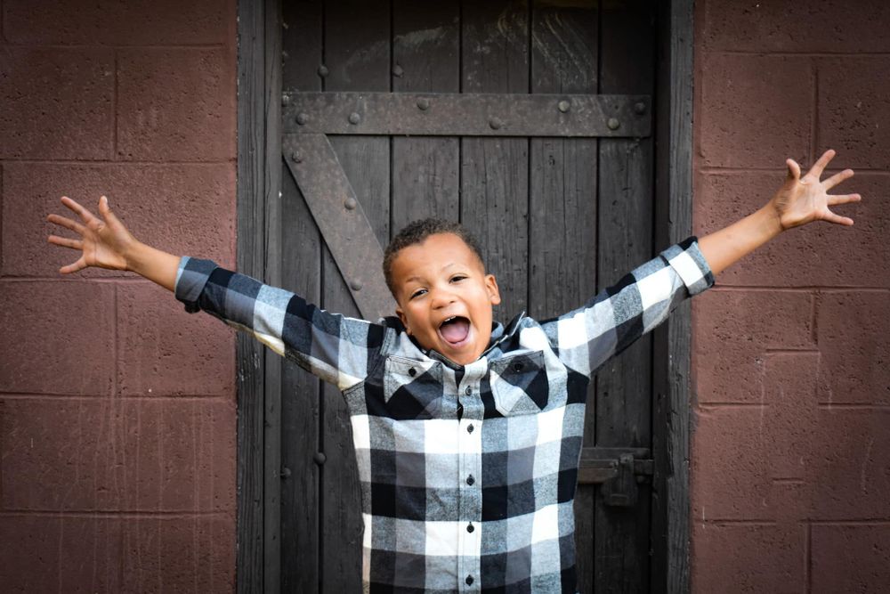 A joyful child in a buffalo plaid shirt spreads their arms wide in front of a wooden door.