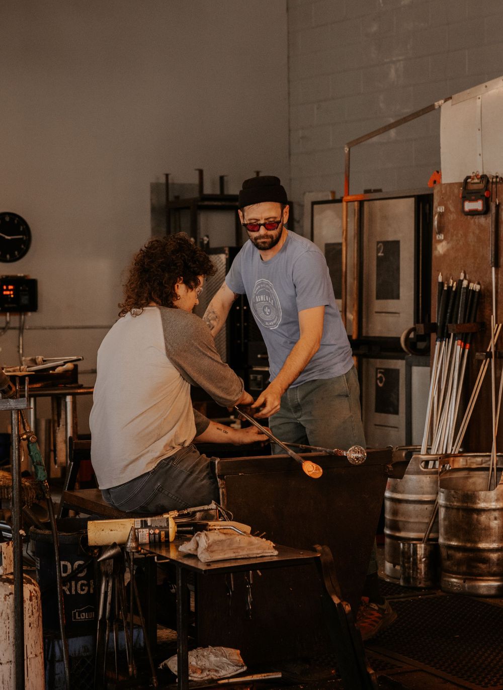 Two artisans work together at a glassblowing workshop, shaping molten glass with traditional tools and equipment.
