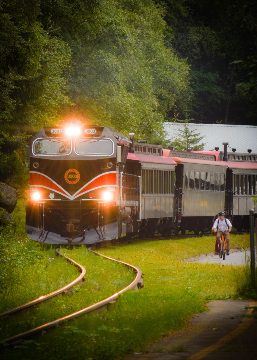 A vintage train with glowing headlights travels along tracks while a cyclist rides nearby.