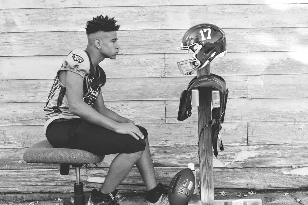 Football player sitting next to helmet on wooden bench in black and white photograph.