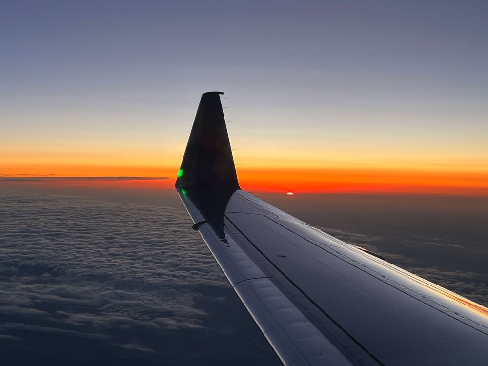 View from airplane window showing wing silhouette against vibrant orange and purple sunset sky over clouds.
