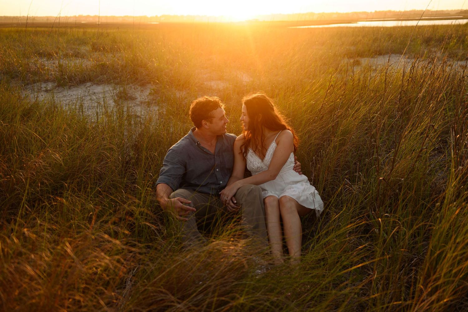 A couple sits together in tall grass at sunset sharing an intimate moment.