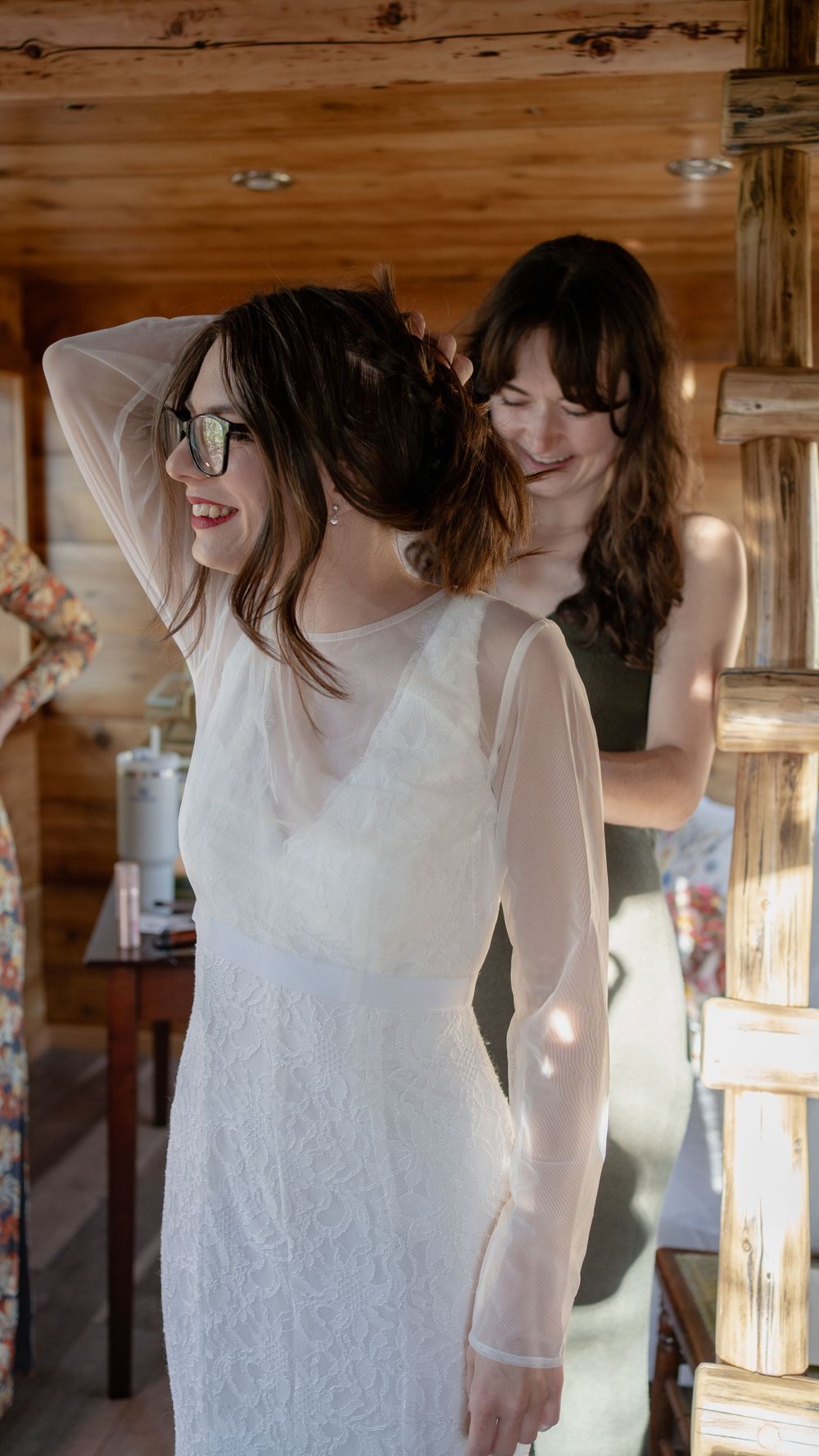 Two people share a joyful moment in a rustic cabin while trying on a white lace wedding dress.