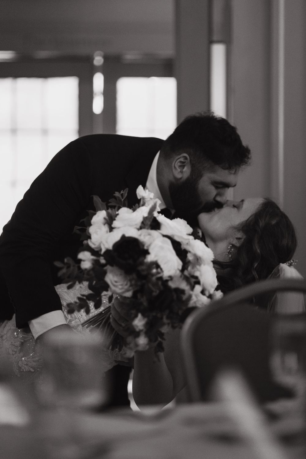 Romantic black and white photo of newlyweds kissing at their reception with floral bouquet.